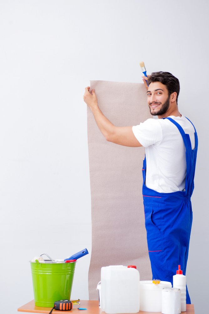 Een rol glasvezelbehang wordt aangebracht op gipsplaten door een persoon met behulp van een behangborstel en andere benodigde gereedschappen.