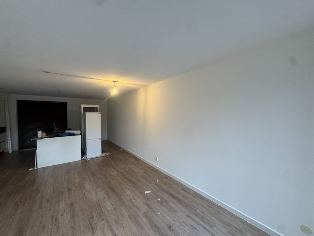 Woonkamer met een laminaatvloer, gladde witte wanden en een keukenblok op de achtergrond