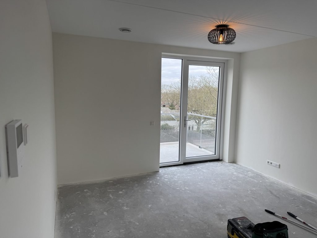 Lege nieuwbouwkamer met strak afgewerkte witte wanden en openslaande deuren naar het balkon